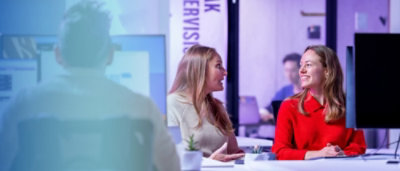 A group of people sitting at a table, with a woman in a red shirt and a man in front of a computer screen.