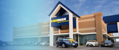 A car dealership with a CARmax sign and cars parked outside under a blue sky.