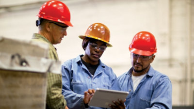 Workers wearing safety helmets reviewing information on a tablet in an industrial setting.