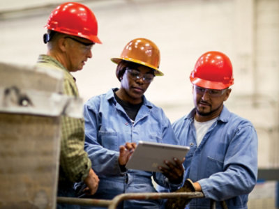 Workers wearing safety helmets reviewing information on a tablet in an industrial setting.