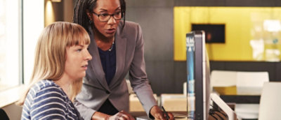 Two colleagues collaborating at a desktop computer while discussing on‑screen content.