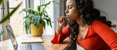 Person working on a laptop at a wooden table near a window with plants.