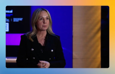 Person speaking on camera in a studio setting, with a play button overlay indicating a video.