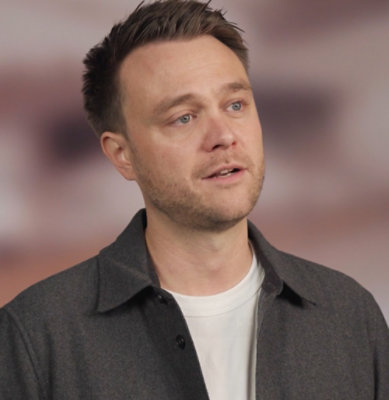 Person speaking on camera in a studio setting with a softly blurred background.