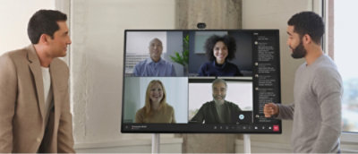 Two men stand by a digital screen displaying a video conference with four participants.