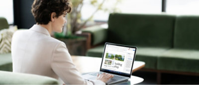 A person with short curly hair, wearing a light-colored blazer, sits using a laptop on a green couch. 