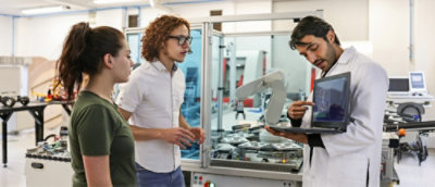 A group of people standing in a room, some looking at a laptop and a machine.