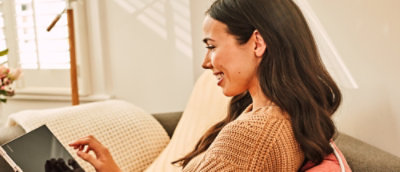 A woman with long dark hair is sitting on a couch, smiling, and using a tablet. 