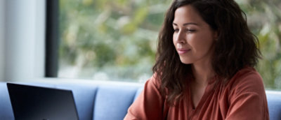 A person with long, dark hair is seated and working on a laptop in a well-lit room with large windows.