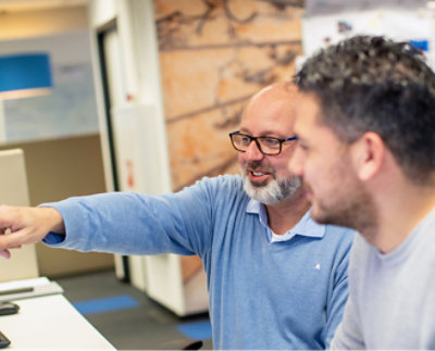 Two men sit at a desk, one wearing glasses and pointing at something on a computer screen.