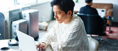 Muž v bílé košili, který pracuje na notebooku u stolu v dobře osvětlené místnosti. V pozadí pracuje další osoba.