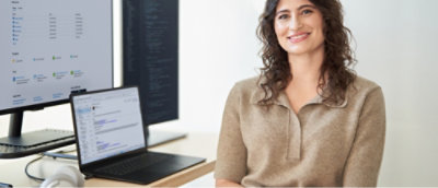 Osoba, která sedí u stolu s notebookem a dvěma monitory zobrazujícími kód a data.