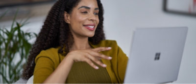 Женщина с вьющимися волосами, в желтом свитере, жестикулирует, глядя на ноутбук.