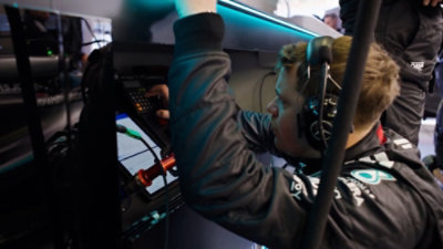 A close-up of a Formula 1 garage workstation shows a crew member wearing a team jacket and a headset.