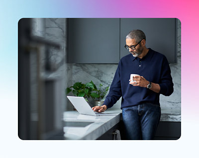 A man stands in a kitchen, holding a mug and looking at a laptop screen on the counter.