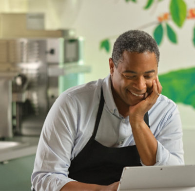 Une personne portant un tablier sourit en regardant une tablette dans une cuisine.