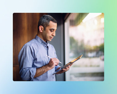 Un homme se tient près d'une fenêtre, tenant et écrivant sur une tablette avec un stylet, vêtu d'une chemise bleue à boutons.