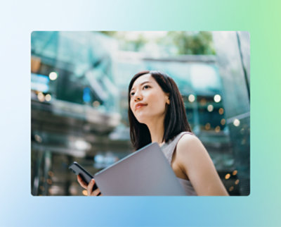 Une femme tient un ordinateur portable et un smartphone, regardant vers le haut, avec un bâtiment moderne en verre en arrière-plan.