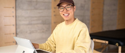 Une personne portant des lunettes et une casquette, assise à une table avec un ordinateur portable, sourit à la caméra.