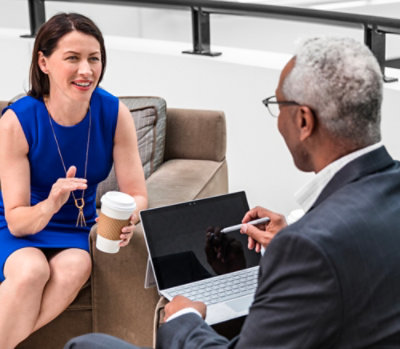 Two professionals seated and discussing work, one using a laptop while the other holds a coffee cup in a casual meeting area.