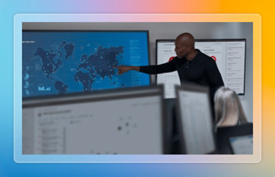 Presenter pointing to analytics and a world map displayed on large screens during a meeting.