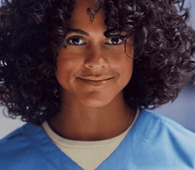Person wearing blue scrubs with curly hair, facing forward in a softly lit indoor setting.