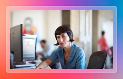 Person seated at a desk using a desktop computer in a shared office environment with others working nearby.
