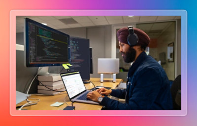 Person working on a laptop at a desk with large monitors displaying code and development tools in an office workspace.