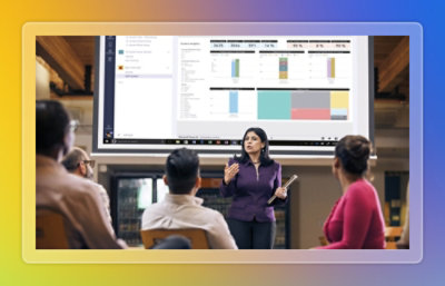 Presenter standing in front of a large screen explaining analytics dashboards and visual reports to a seated audience.