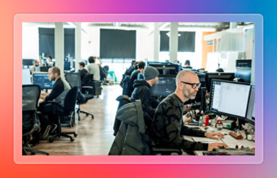 Image of a modern open office where multiple people work at desktop computers in a collaborative development environment.