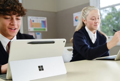 Two students seated at a desk using a Microsoft Surface device and a tablet while working on a classroom assignment.
