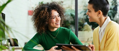 Zwei Personen lächeln sich in einem hellen Raum an und halten jeweils ein Tablet in der Hand – die eine trägt einen grünen Pullover, die andere eine gelbe Strickjacke.