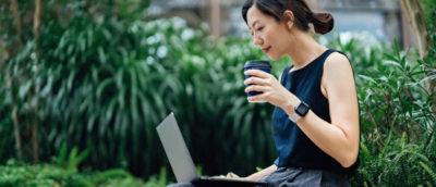 Person using a laptop outdoors while holding a coffee cup.