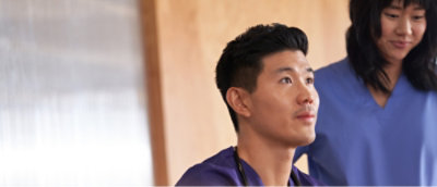 A man in a purple shirt looks ahead thoughtfully while a woman in a blue shirt stands beside him, smiling slightly.