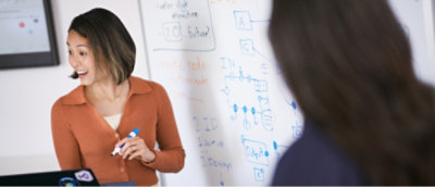 A woman holding a blue marker talks and smiles while standing near a whiteboard filled with various diagrams and notes.