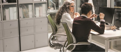Two people sit at a desk in an office, examining a computer screen together. 
