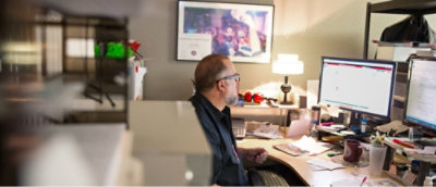 A person with glasses sits at a cluttered desk, working on a computer.