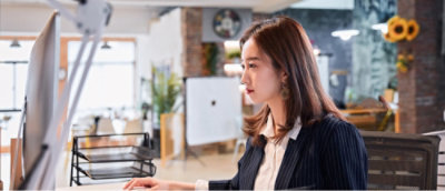 A woman sits at a desk in an office, focused on her computer screen.
