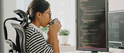 A person wearing glasses and a striped shirt sits at a desk drinking from a mug while looking at two monitors displaying code.
