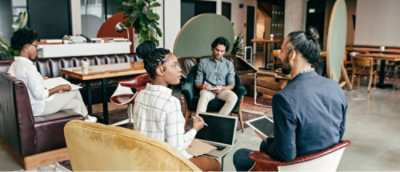 Group of four people in a modern office seating area engaged in conversation, some holding laptops and notepads.