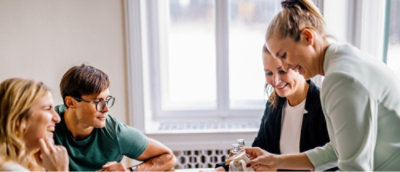 Four people sitting at a table, engaged in a discussion, with one person standing and showing something to the group.