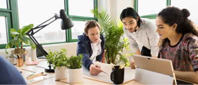 Three people collaborate at a desk with plants, a lamp, and a tablet.