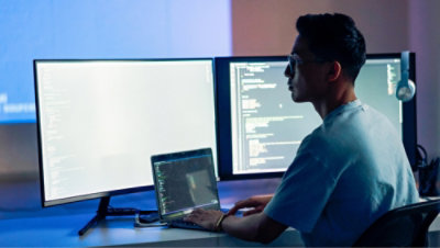 A person works at a desk with three monitors displaying code and a laptop.