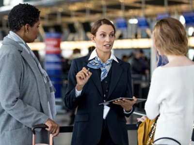 A uniformed airline employee speaks to two travelers at an airport, holding a tablet and gesturing with her hand.