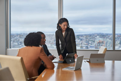 Three people in a conference room work on laptops, with one standing and pointing at a screen.