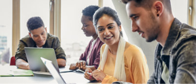 Four people sitting at a table with laptops, engaged in a discussion, in a bright room with large windows.