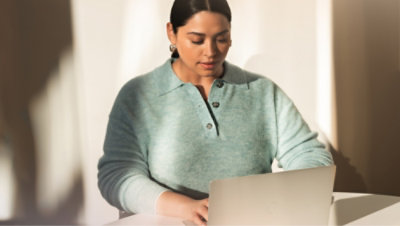 A person in a teal sweater is seated at a white table using a laptop.