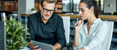 Two people are seated at a desk, discussing something on a tablet. 
