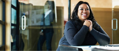 A smiling woman sits at a desk in a modern office. In the background, a person is visible through a glass wall.