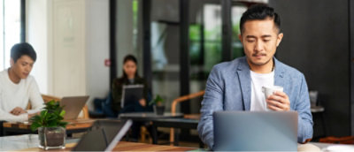 People sitting at desks in a workspace. The man in the foreground is holding a mug and looking at a laptop.